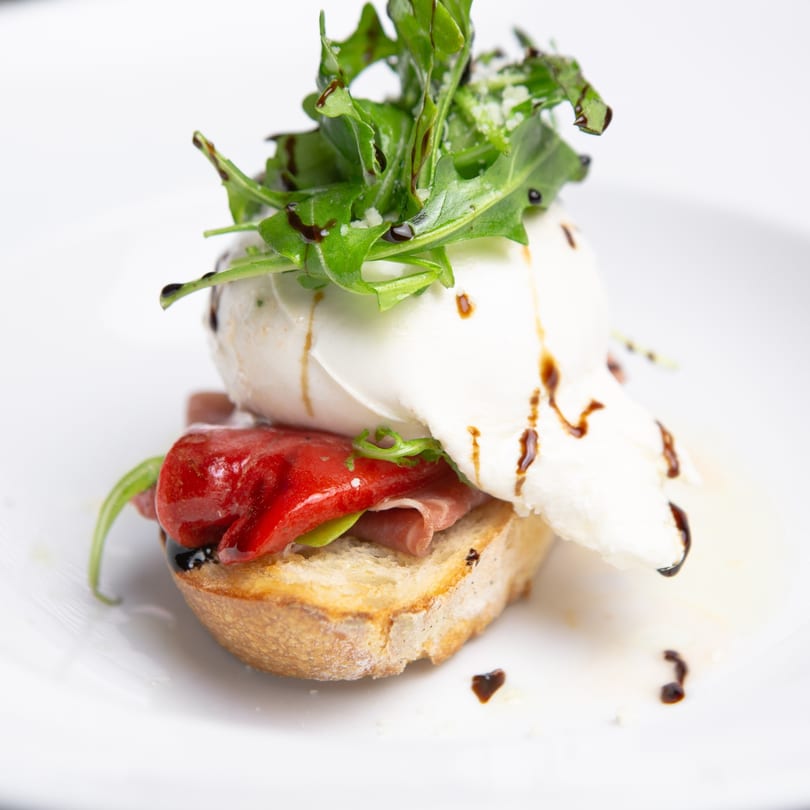 Italian bruschetta with burrata, roasted red peppers, prosciutto, arugula, and balsamic glaze on a white plate.