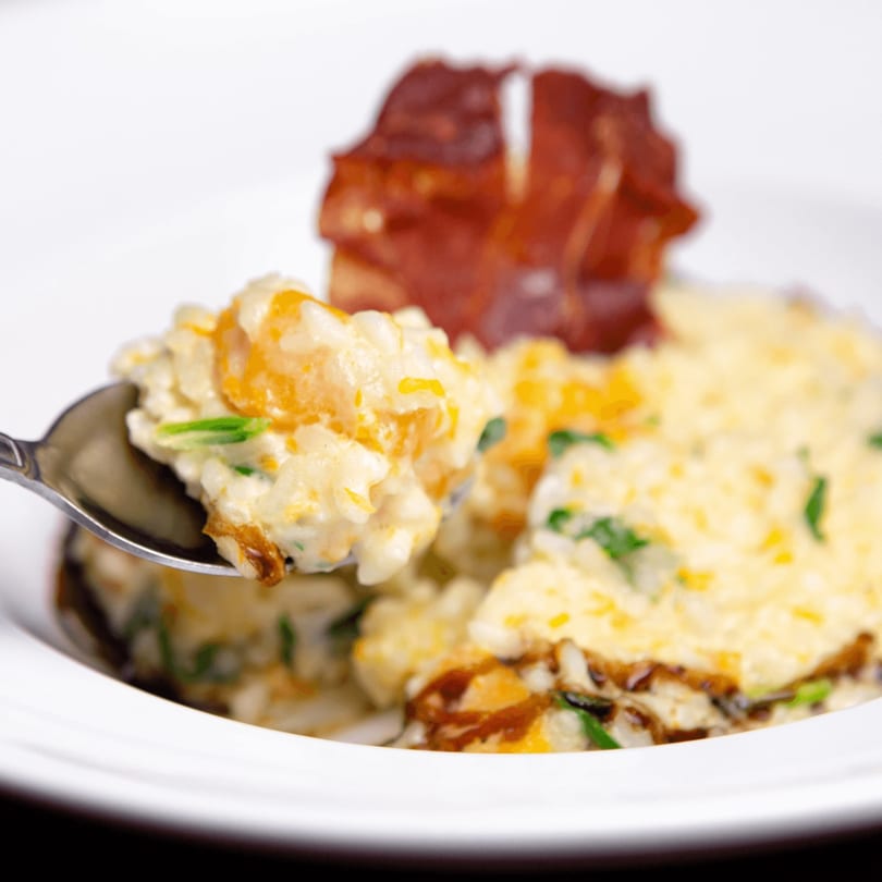 Creamy risotto with cheese and green herbs, with a spoonful being lifted, and crispy bacon in the background.