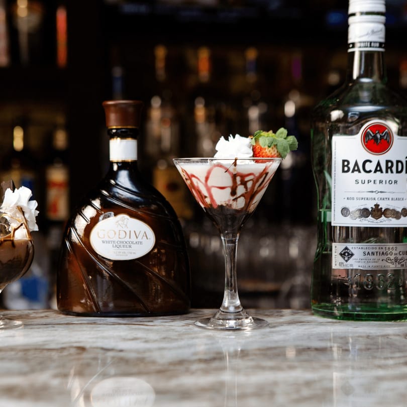 A martini glass with a chocolate-drizzled drink, whipped cream, and strawberry, flanked by liquor bottles.