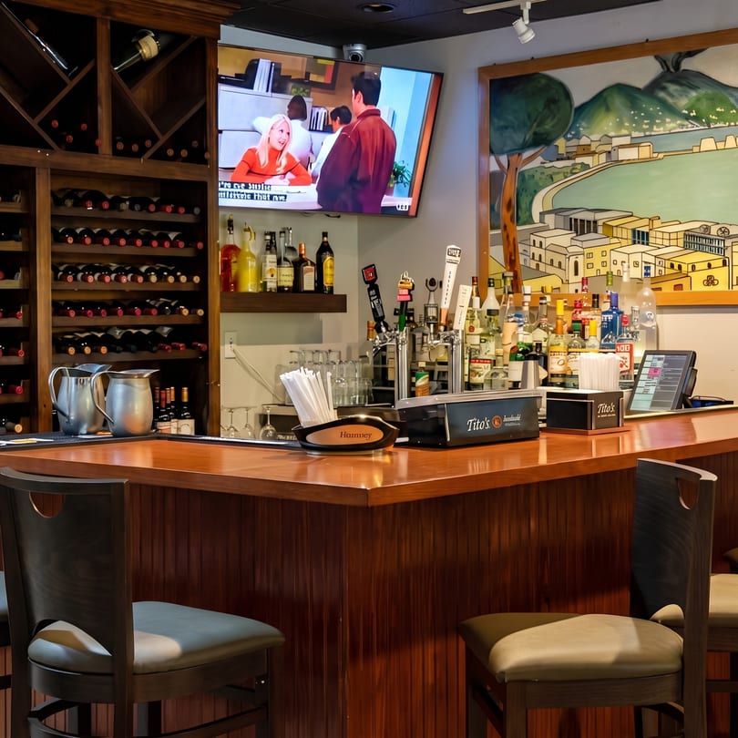 A well-stocked bar with a wooden counter, wine rack, liquor bottles, beer taps, and a TV playing a show.