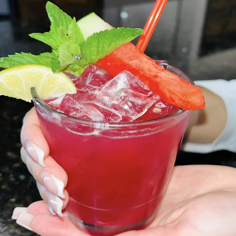 A vibrant red cocktail with ice, garnished with mint, lemon, and watermelon, held by a hand.